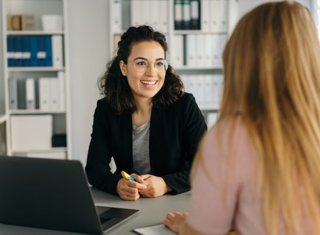 Two young women having a business meeting in the office with over the shoulder focus to a smart you woman sitting listening with a smile