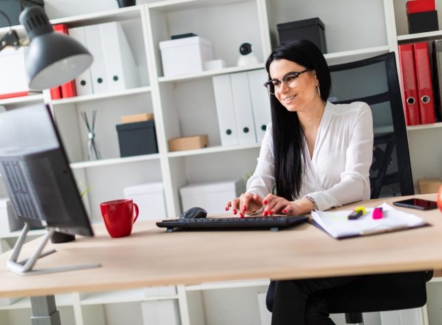 A young girl with glasses, wearing a white blouse and black trousers, works in the office. The girl has long straight black hair. photo with depth of field