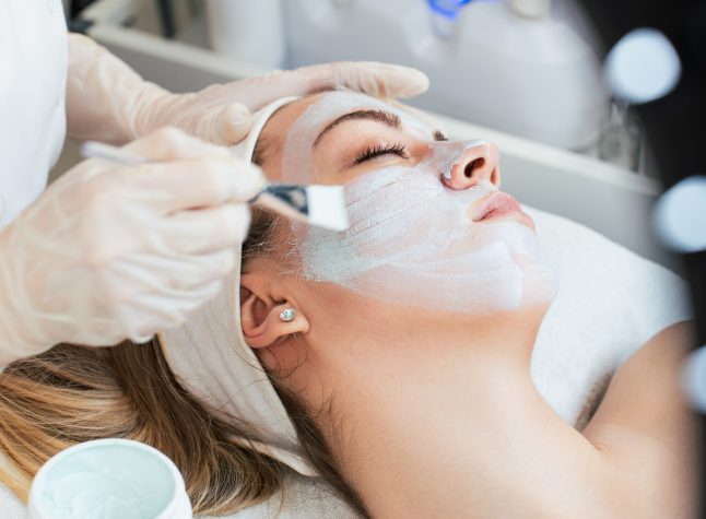 Beautiful and attractive young adult woman receiving professional beauty or skincare treatment with herbal clay mask. Modern and popular facial care treatment concept. Spa salon. High angle view.